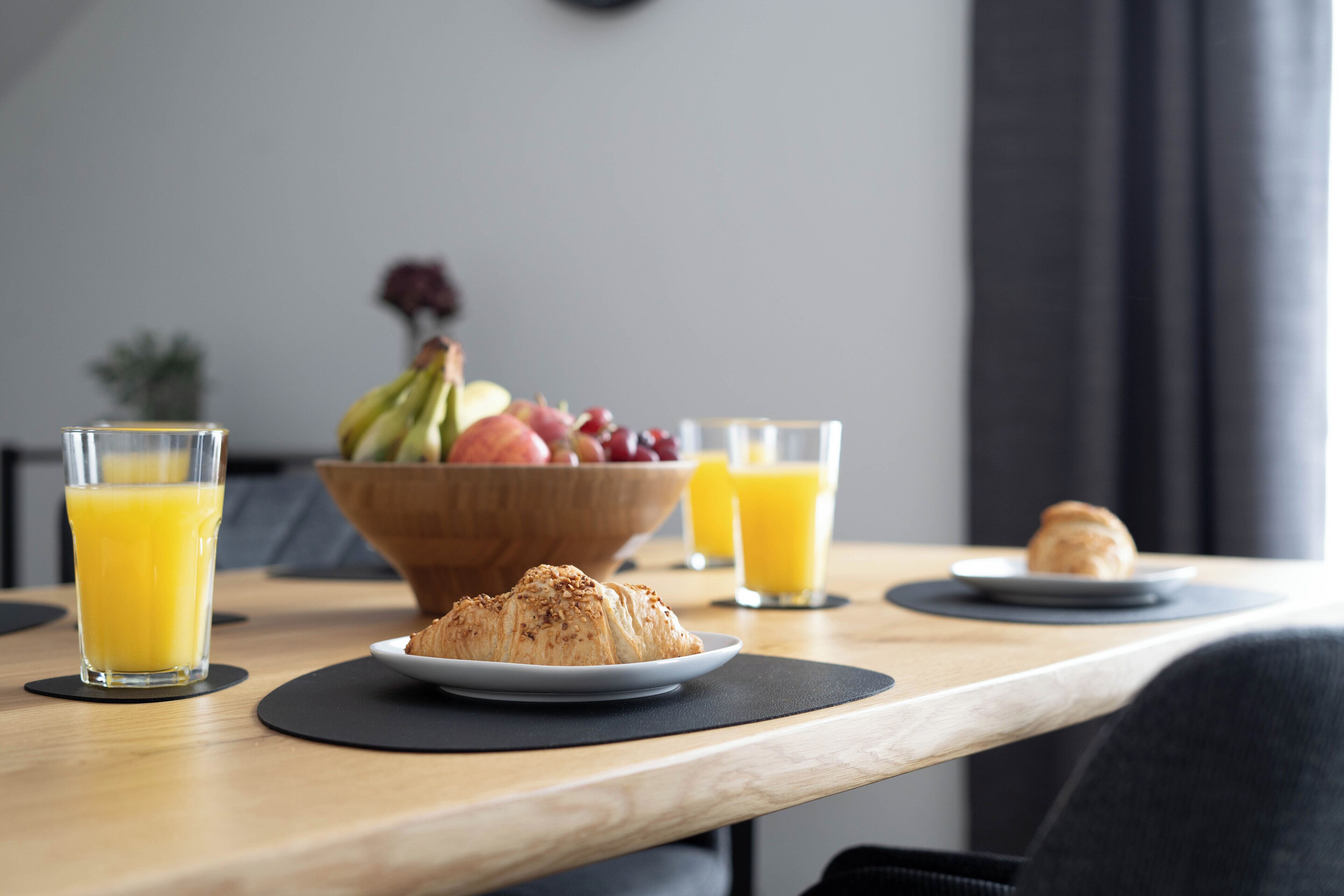 Aamiaispöydällä on croissantteja, hedelmäkulho ja lasit täynnä appelsiinimehua.
