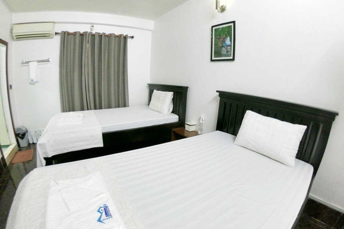 Habitación de hotel con dos camas, una ventana con cortinas, un equipo de aire acondicionado, un cuadro en la pared y una mesita con una botella.