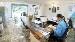 A room with a desk, computer, and various office supplies. There is a window with a view of trees and a door leading to another room. - Sarah Guest House (Siem Reap)