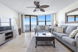 A spacious living room with a large flat-screen TV, a ceiling fan, a sofa set, and a view of the ocean through large windows. - Lighthouse Shores Condo 403 (Ponce Inlet)