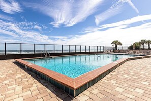 A rectangular swimming pool with a tiled edge, surrounded by a brick deck, and a metal fence with a view of the ocean. - Oceanfront Condo on traffic free beach - Great views from 403 Lighthouse Shores (Ponce Inlet)