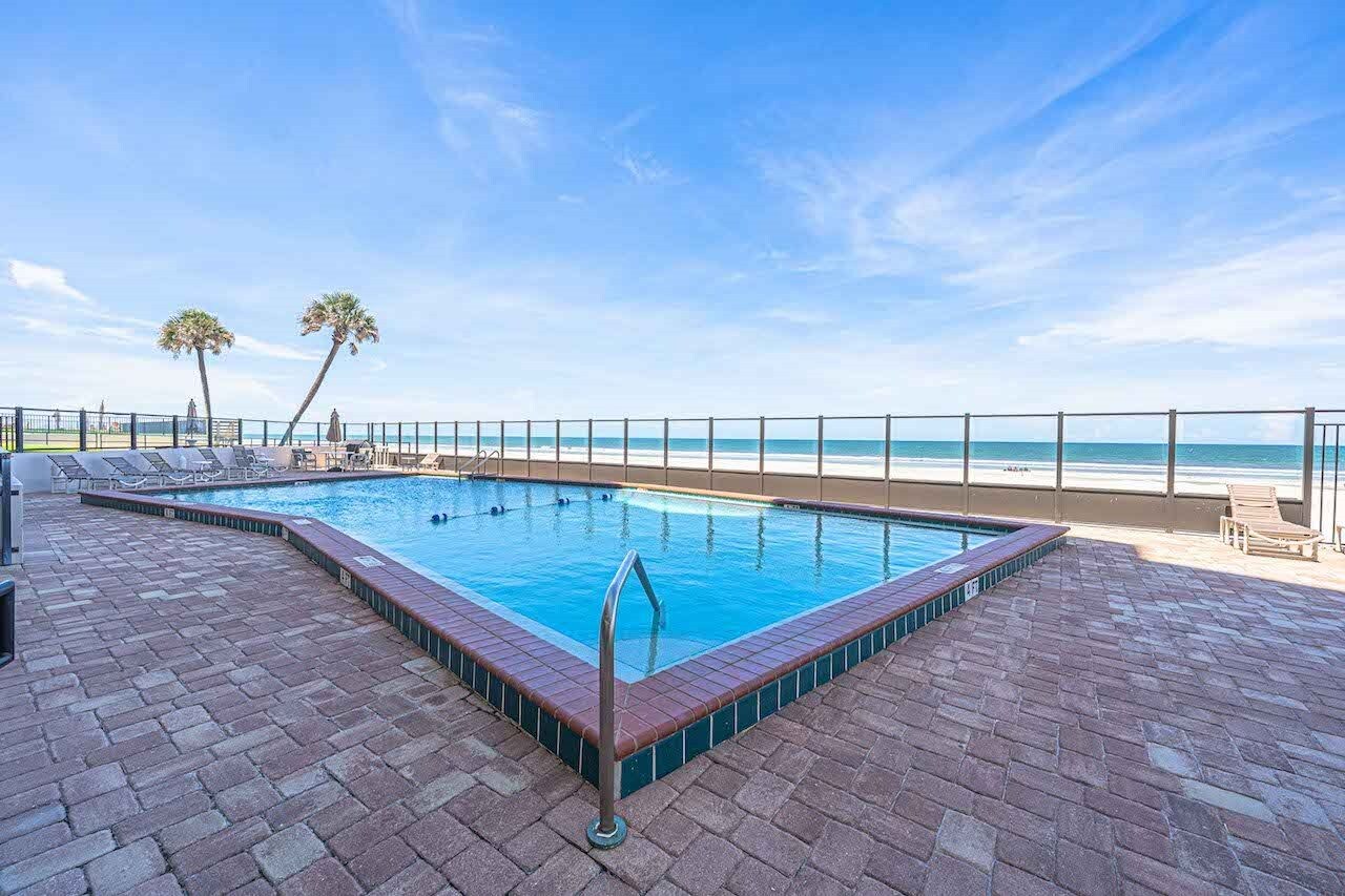 Piscina rectangular con escalera metálica, rodeada de un patio de ladrillo, con vistas a una playa y un cielo despejado.