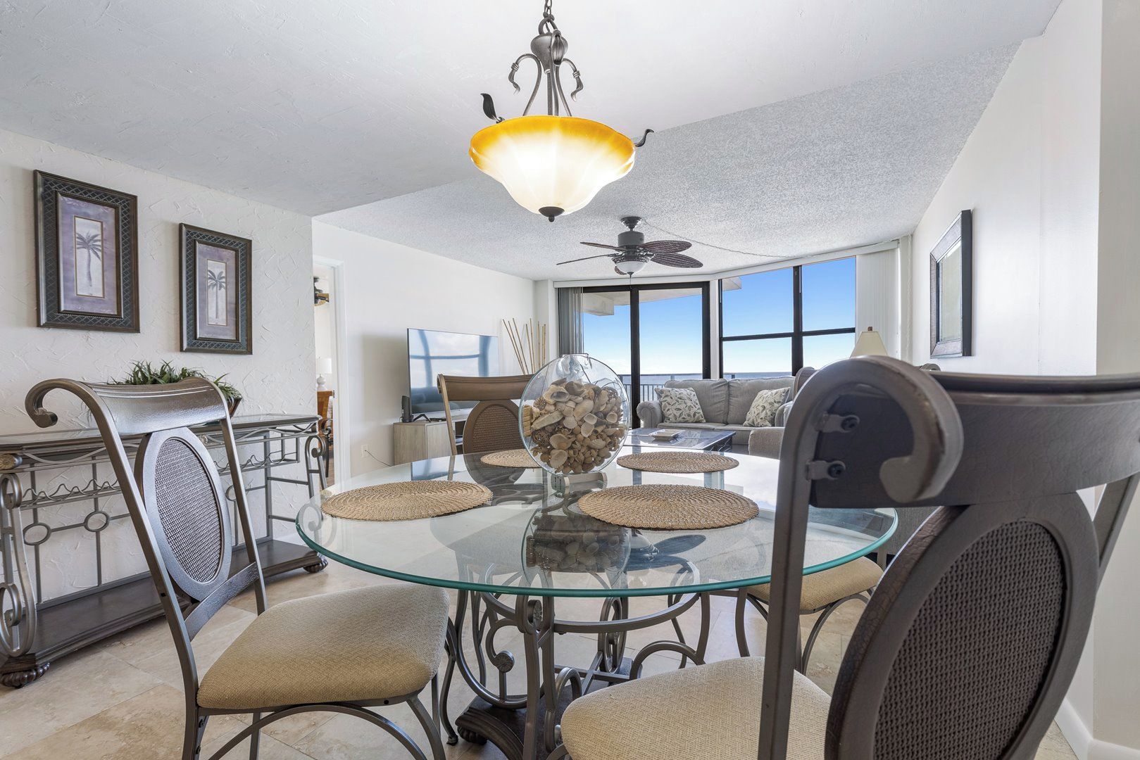A dining area with a glass table, chairs, a chandelier, and a view of the outdoors.