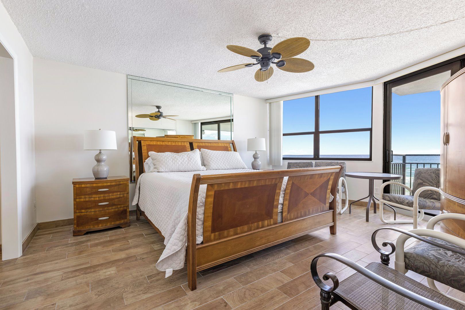 A hotel room with a wooden bed, a ceiling fan, a large window, and a balcony with a view of the ocean.