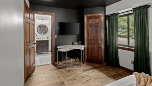 Una stanza con pavimento in legno, uno specchio rotondo, un tavolino con una sedia, una televisione a muro e una finestra con tende.