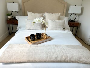 Una cama bien tendida con una bandeja de tazas de café, un florero con flores y un suave plaid blanco.