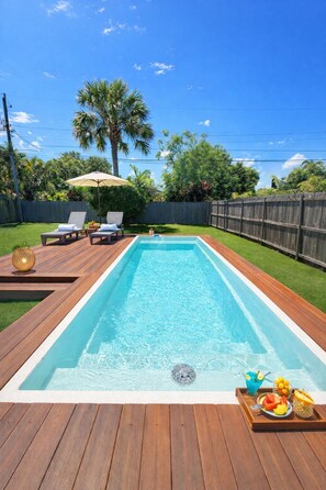 Una piscina rectangular con maderas, sillas de descanso y una mesa con fruta y bebidas.