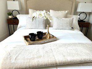 Un lit bien fait, recouvert d’une couette blanche, deux tasses à café noires ornées d’inscriptions, un vase doré et des orchidées blanches.