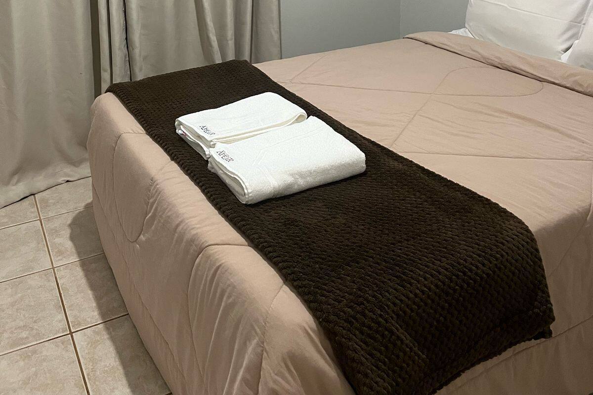 A neatly made bed with a beige bedspread and a brown blanket, a white pillow, and a folded white towel on the bed.