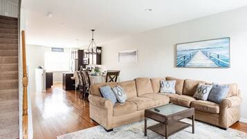 A spacious living room with a beige sectional sofa, a glass coffee table, and a dining area with a table and chairs.