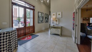 Un vestíbulo de hotel con una puerta de madera grande, una araña, una mesa a cuadros de galgo, una mesa blanca pequeña y cuadros en la pared.