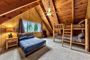 A cozy attic bedroom with a bunk bed, a single bed, a window with a view of trees, and a ceiling fan.