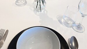 Un tavolo da pranzo con un vaso bianco contenente fiori bianchi e un ramo decorativo, insieme a una ciotola e posate.