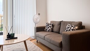 Un soggiorno moderno con un divano grigio, cuscini decorati, una ventola a piantana, un tavolino rotondo e una lampada da terra.