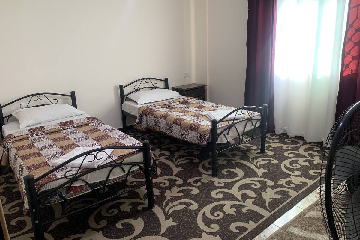 A room with two single beds, a small table, a chair, and a patterned carpet.