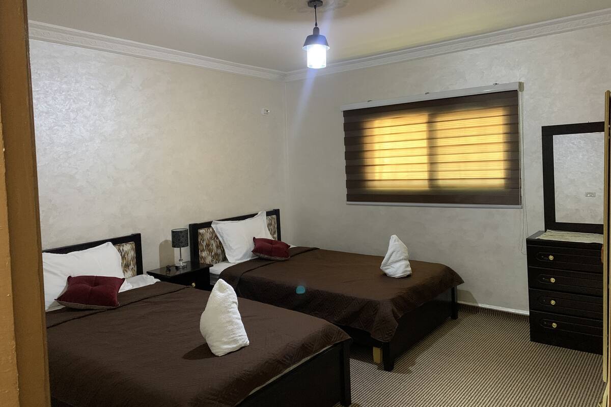 A hotel room with two beds, a nightstand, a dresser, a window with blinds, and a ceiling light.