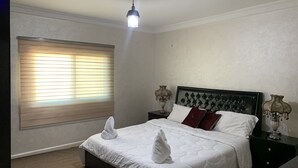 A neatly made bed with white linens and two towels placed on it. There is a window with blinds, a bedside lamp, and a decorative wall piece on the nightstand. - Paradise Tree  (Wadi Musa)