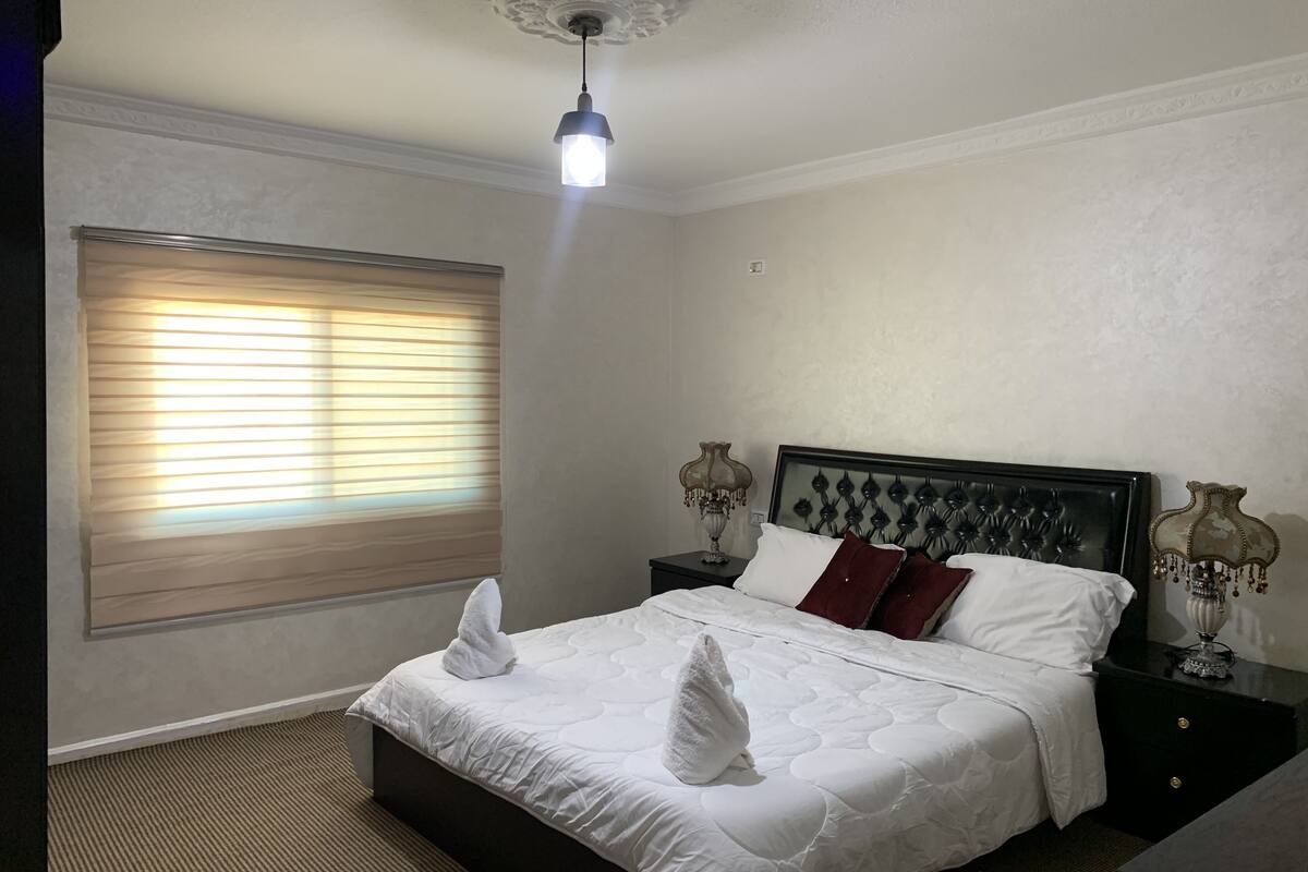 A neatly made bed with white linens and two towels placed on it. There is a window with blinds, a bedside lamp, and a decorative wall piece on the nightstand.