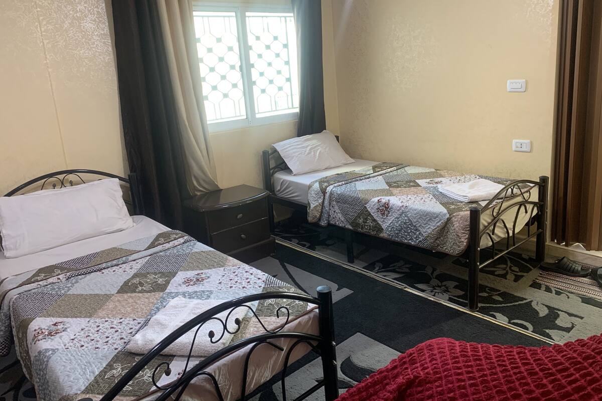 A small, cozy room with two single beds, a nightstand, a window with a view, and a red blanket on the floor.