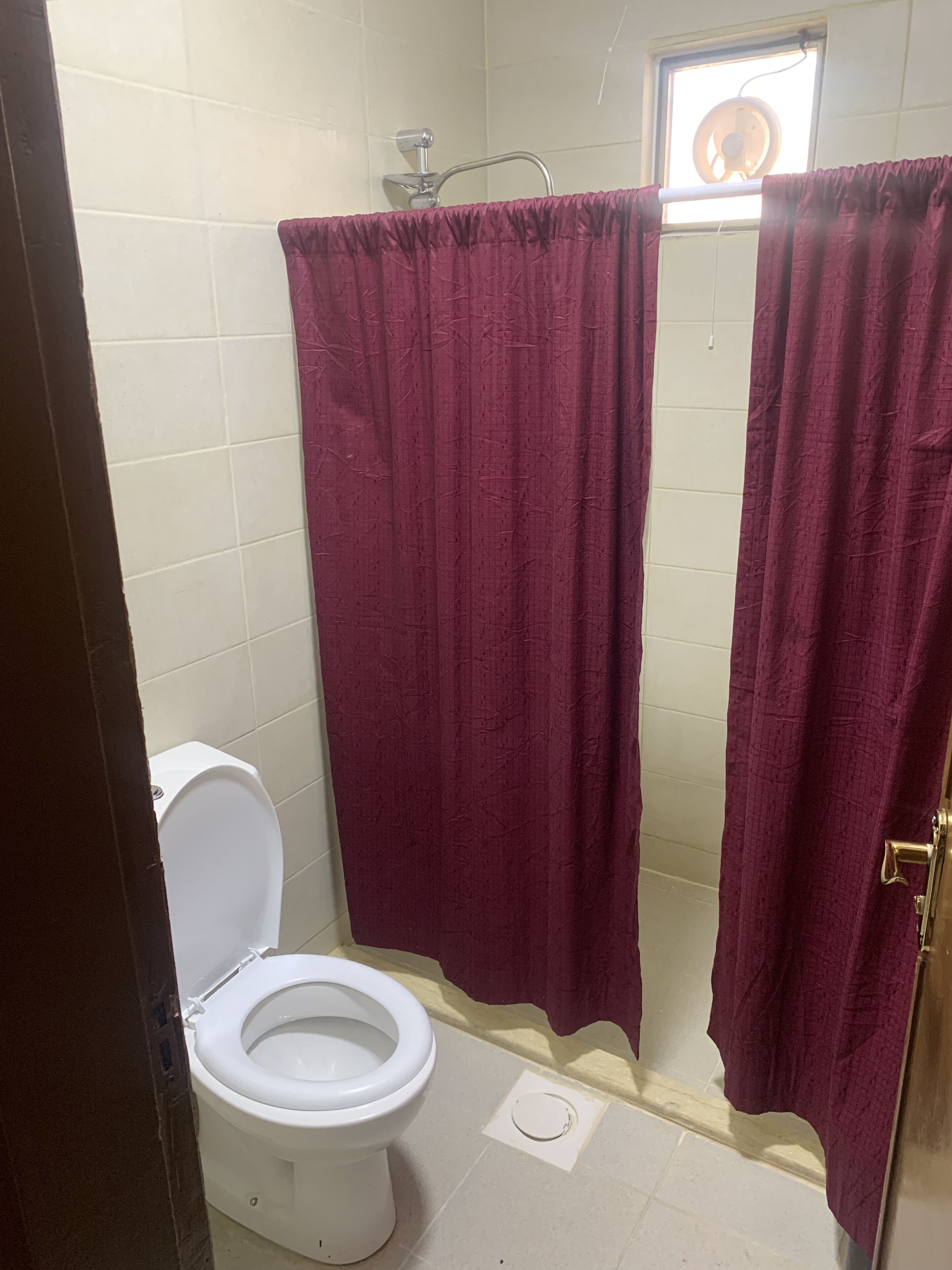 A bathroom with a white toilet, a shower curtain, and a window with a fan.