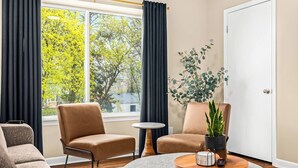 A modern living room with two leather armchairs, a round coffee table with a plant, and a view of trees through a large window.