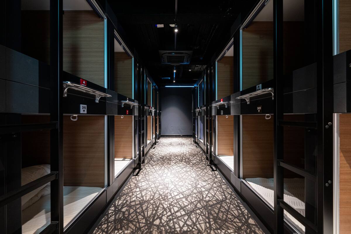 A hotel corridor with bunk beds, a patterned carpet, and a dark ceiling with lighting fixtures.