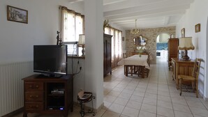 A spacious living area with a television, dining table, and chairs. A kitchen is visible in the background. - Maison de Campagne (Saint-Amans-Valtoret)