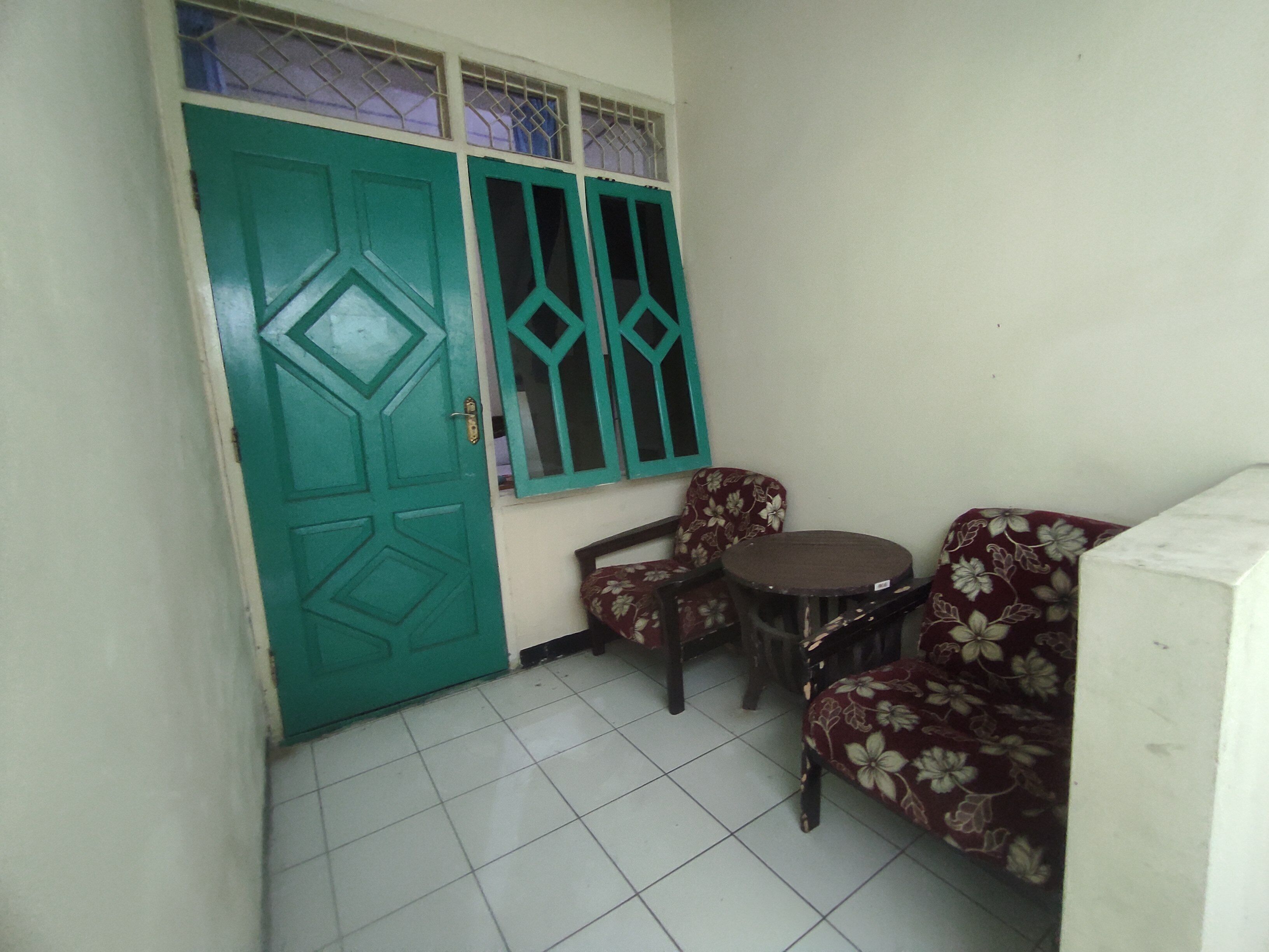 A small room with a green door, a window with metal bars, two floral-patterned chairs, and a small round table.
