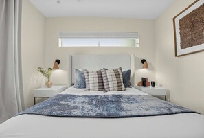 Une chambre à coucher avec un grand lit, deux tables de chevet avec des lampes, une fenêtre avec des stores et une œuvre d’art accrochée au mur.