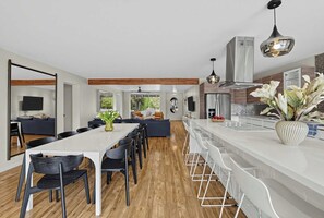 A modern kitchen with a long countertop, a dining area with a white table and black chairs, and a living area with a sofa and chairs. - Indigo Oasis, Pool, Sleeps 18 (Phoenix)