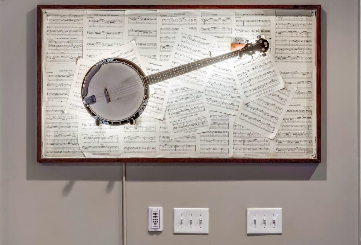 A framed display of a banjo on sheet music, with wall-mounted electrical outlets below.