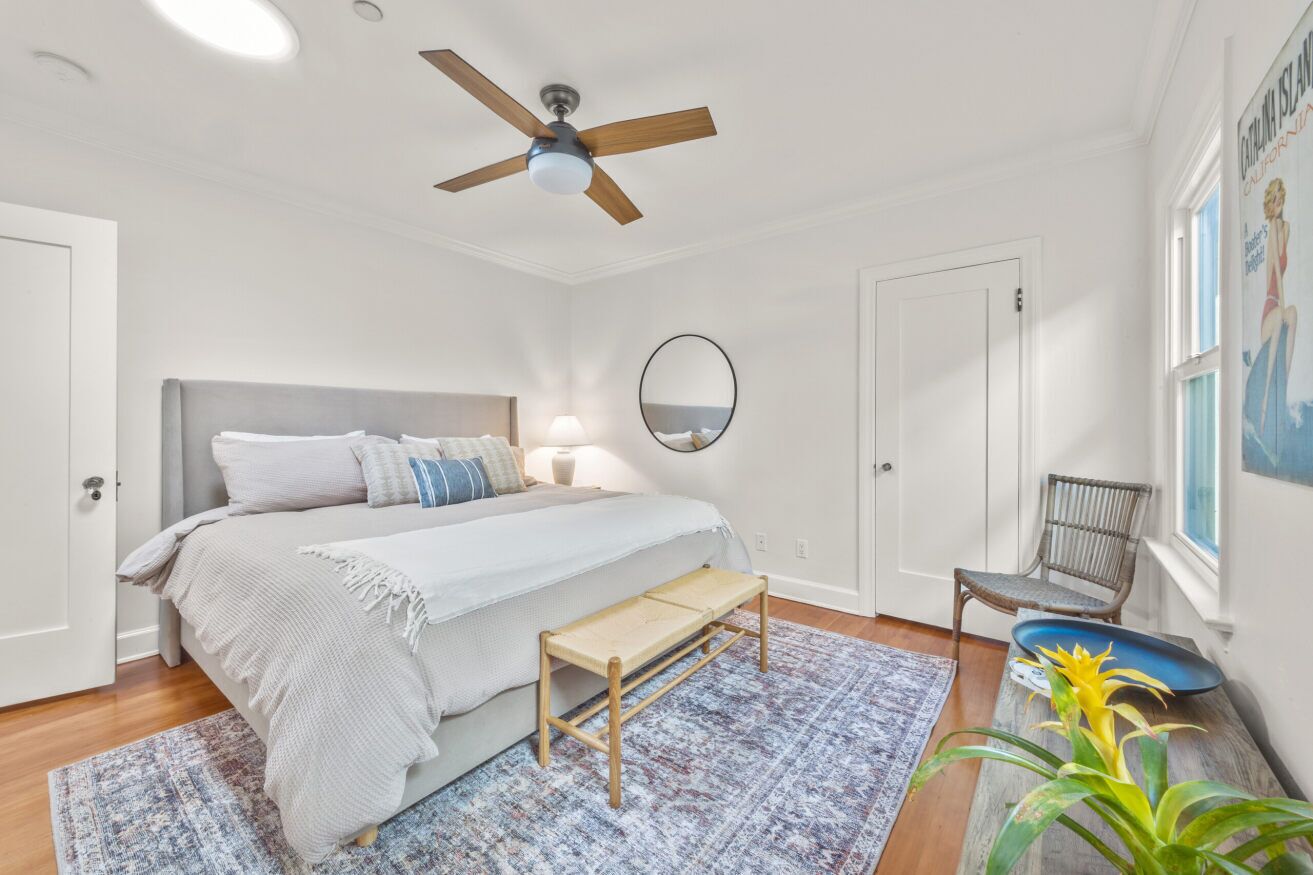 Een slaapkamer met een bed, een plafondventilator, een nachtkastje met een lamp, een spiegel, een stoel en een raam met uitzicht op de oceaan.