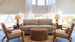Ein gemütliches Wohnzimmer mit einer Couch, zwei Sesseln, einem gewebten Hocker und zwei Lampen.