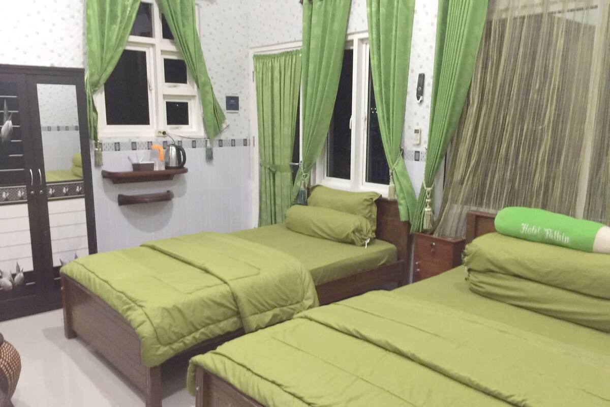 Een kamer met twee enkele bedden, elk met groene beddengoed, een houten hoofdbord en een klein nachtkastje. Er is een raam met groene gordijnen, een spiegel en een plankje met een waterkoker en andere spullen.