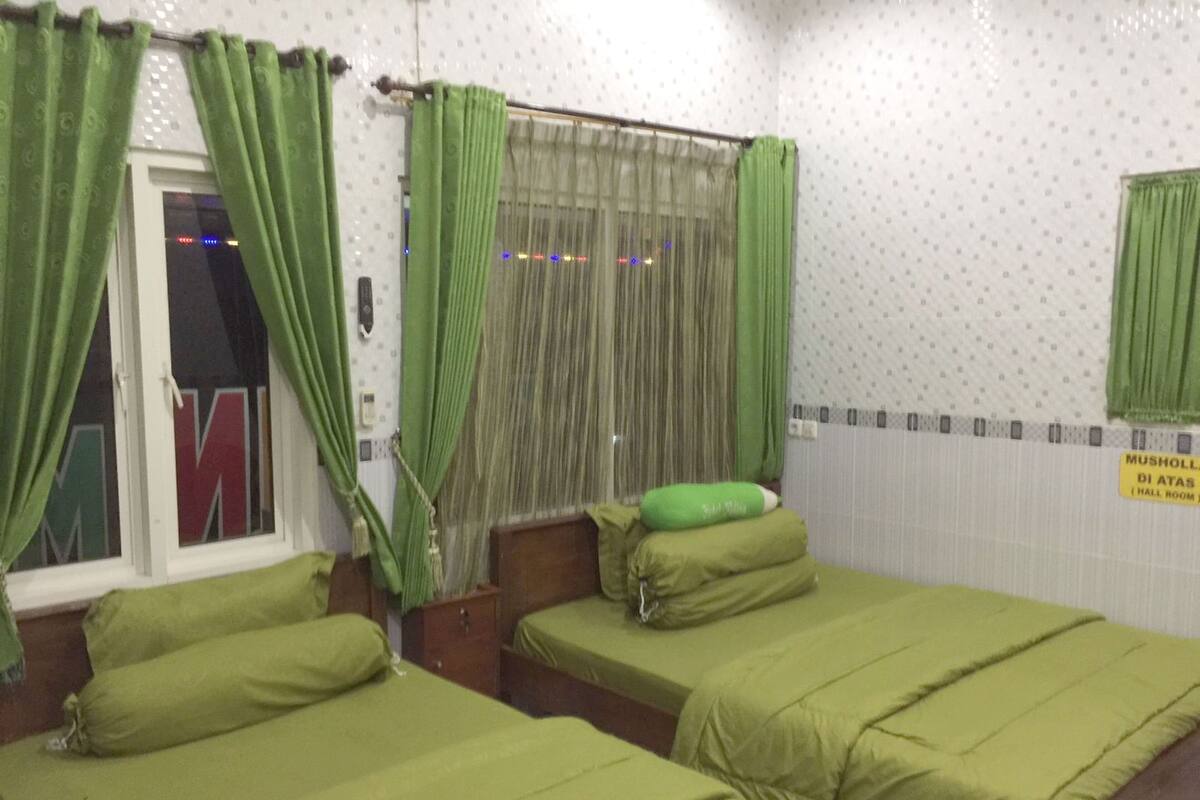 Een kamer met twee bedden, groen beddengoed, een raam met groene gordijnen en een bordje met de tekst 'hal kamer'.