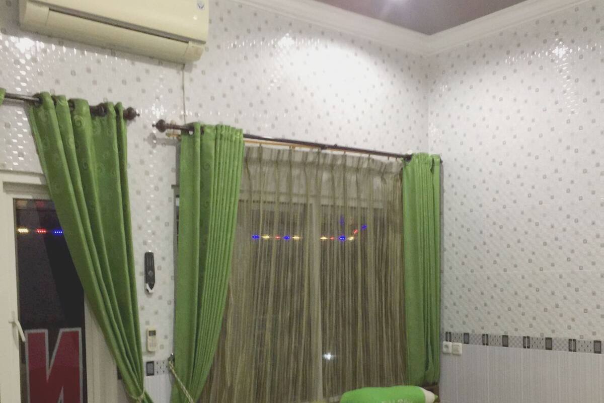 Een kamer met twee bedden, groen beddengoed, een raam met groene gordijnen en een airconditioning op de muur.