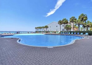 A swimming pool with lounge chairs and palm trees, a building in the background, and a clear sky.