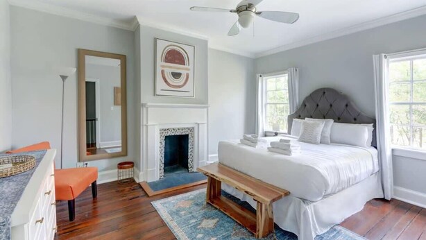 A bedroom with a bed, a bench, a fireplace, a ceiling fan, and a large window. - The Tea Olive - A, Fresh | Updated | Dog-Friendly (Savannah)