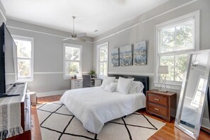A bedroom with a large bed, a desk, and a chair. There are three windows and a ceiling fan.
