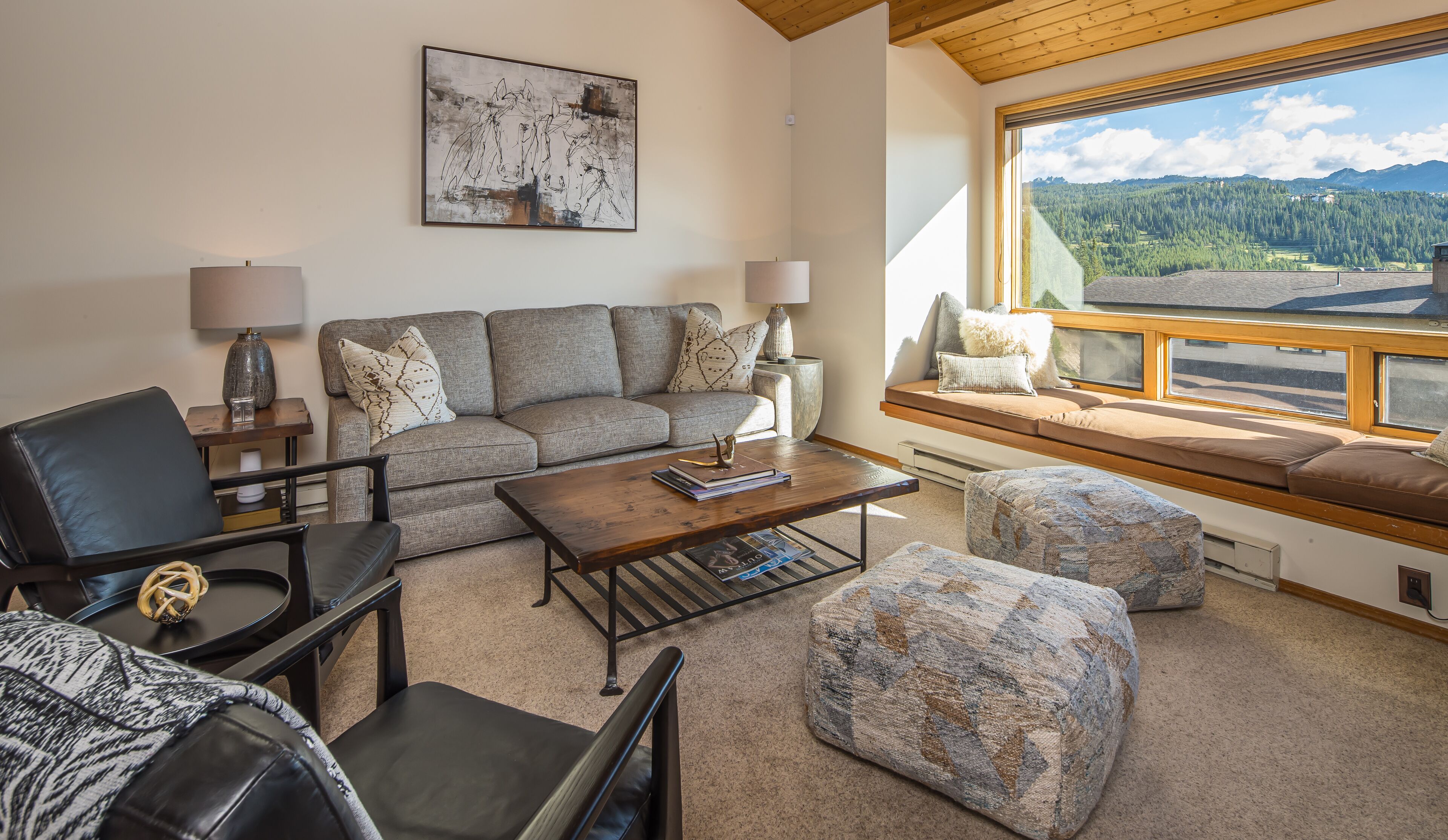 A cozy living room with a sofa, coffee table, and ottoman, a painting on the wall, and a large window with a mountain view.