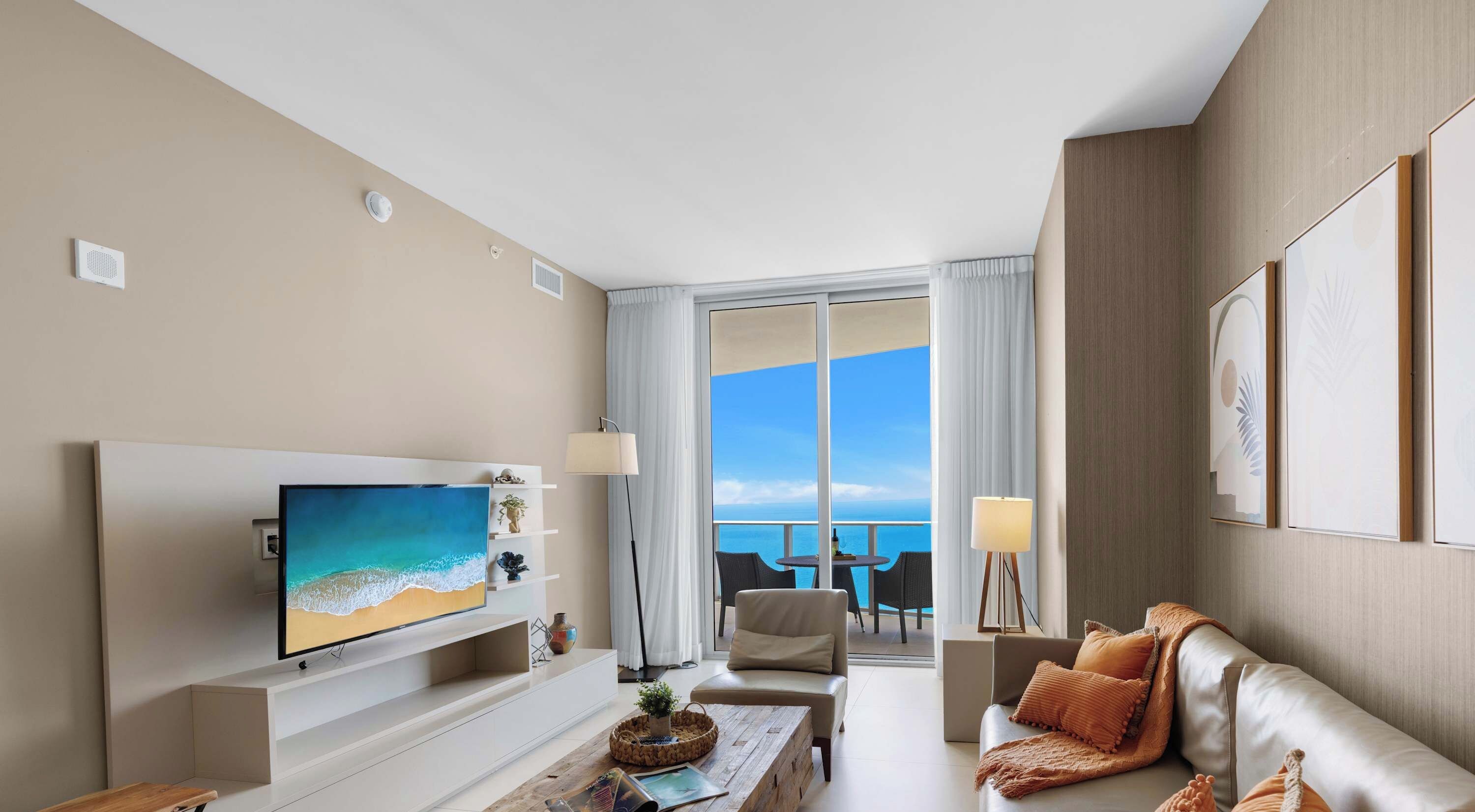 Habitación de hotel moderna con un televisor grande, asientos cómodos, un balcón con vistas al mar y un ambiente bien iluminado.