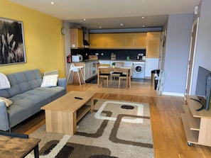 A modern living room with a sofa, coffee table, and a rug. A kitchen with wooden cabinets and appliances is visible in the background. - 12 Courthouse Square Apt with Parking (Clifden)