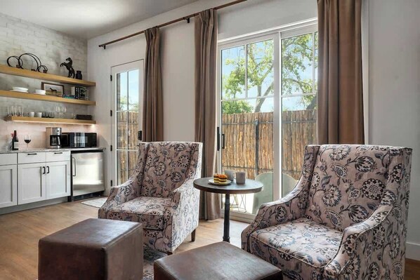 A modern living room with two patterned armchairs, a small round table, and a kitchen area with white cabinets and a coffee machine.