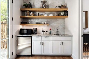 A compact kitchen with white cabinets, a stainless steel dishwasher, a microwave, and a coffee maker. There are wooden shelves above the counter holding various kitchen items.
