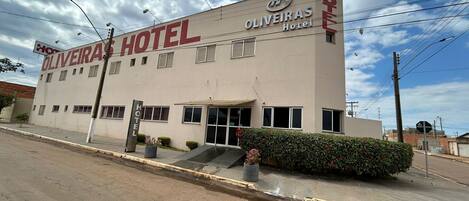 The exterior of the Oliveira Hotel, featuring a simple facade with a prominent sign, a small entrance with steps, and a parking lot in front.