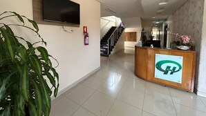 Zona de recepción de hotel con un televisor de pantalla plana montado en la pared, mostrador de recepción con un logotipo, extintor y una escalera que conduce a otro piso.