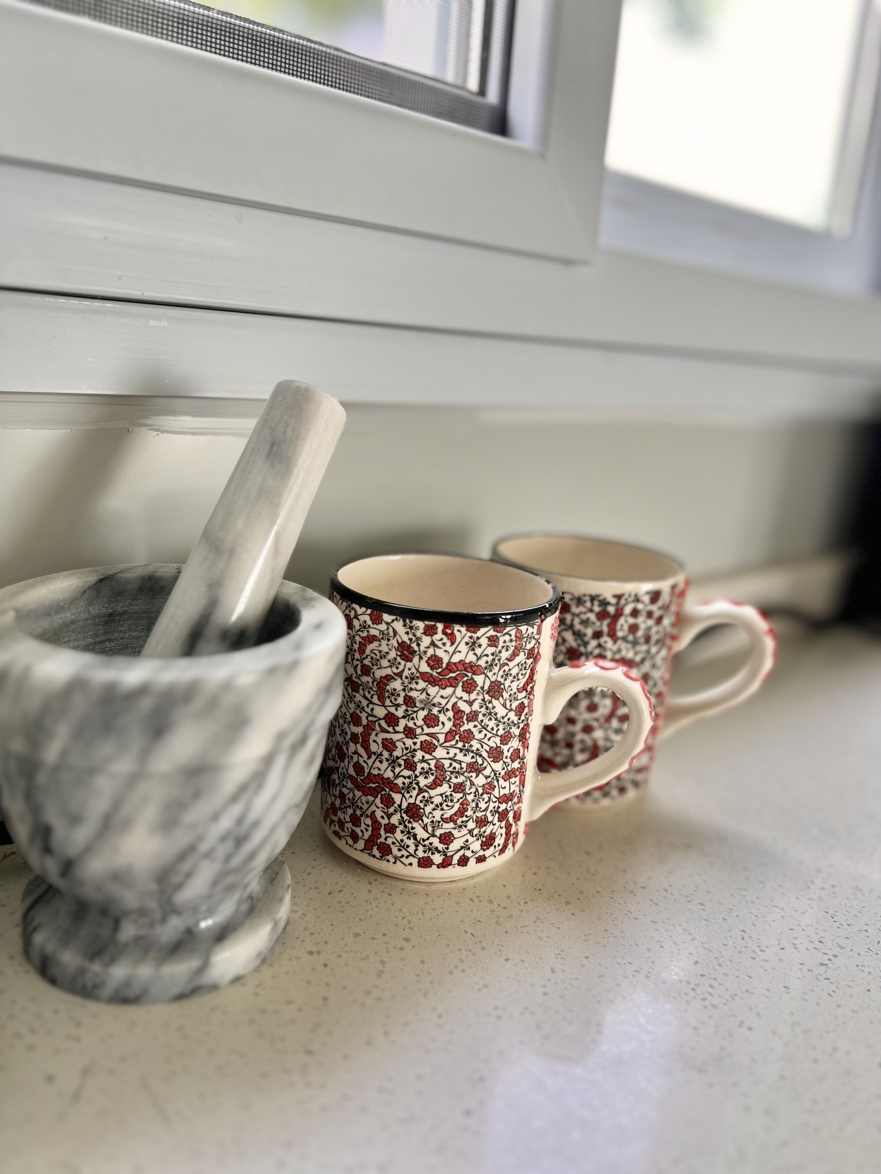 Un mortero y pilón de mármol, y dos tazas estampadas sobre una encimera cerca de una ventana.