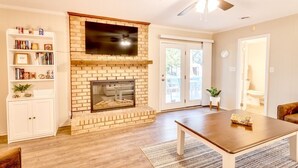 A living room with a brick fireplace, a flat-screen TV mounted above it, a ceiling fan, and a wooden coffee table. - Spacious Retreat Near Lake Hartwell, Clemson & Greenville! (Anderson)
