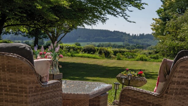 A wicker table with a vase of flowers and a potted plant, set outdoors with a view of a green landscape and trees. - Grand Historical Villa With Stunning Garden (Ross-on-Wye)