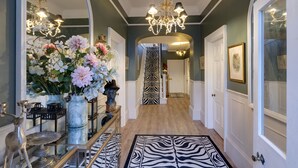 A hallway with a zebra-patterned rug, a chandelier, and a mirror reflecting flowers and a bust. - Grand Historical Villa With Stunning Garden (Ross-on-Wye)
